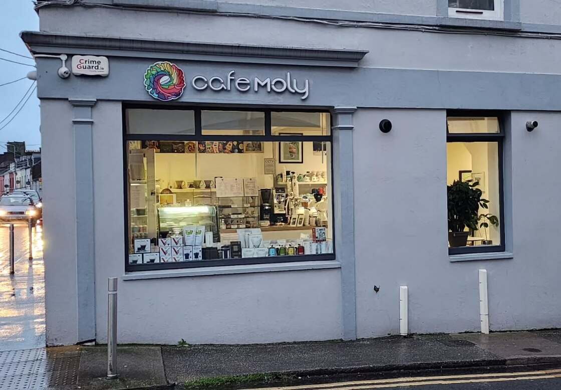 The corner shopfront of Cafe Moly at dusk, grey-blue paint, the rainbow roundel and 'cafe moly' sign above a lit window.