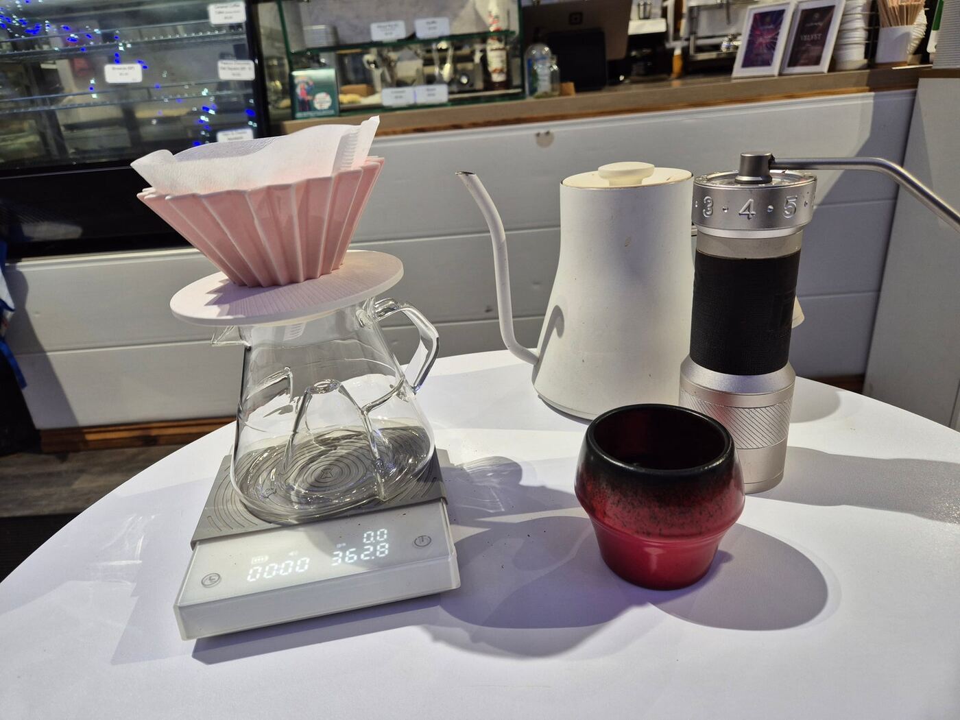 A pour-over set on a white table: Origami dripper, glass server, scale, kettle, hand grinder and a red-black cup.